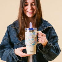 A smiling woman in a denim jacket holds the-darling-effect’s Secret Garden Blue 20 oz Lifestyle Flip Straw Tumbler, featuring a floral pattern, leak-resistant lid, and straw, with "The Thirsty Effect" text against a light beige background.