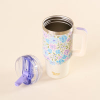 The Secret Garden Blue 20 oz Lifestyle Flip Straw Tumbler from the-darling-effect, with a handle and pastel floral design, sits on a cream surface. Its purple and clear lid is beside it, and “the smiLing effect” is printed in gold on the tumbler.