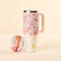 The Secret Garden Pink 40 oz Lifestyle Flip Straw Tumbler by the-darling-effect, featuring a colorful floral pattern, a leak-resistant clear lid with pink straw, and yellow branding, sits on a light surface with its handle visible.