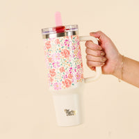 A hand holds the-darling-effect Secret Garden Pink 40 oz Lifestyle Flip Straw Tumbler with a floral pattern, handle, clear leak-resistant lid, red flip straw, and logo on the cream-colored bottom section.