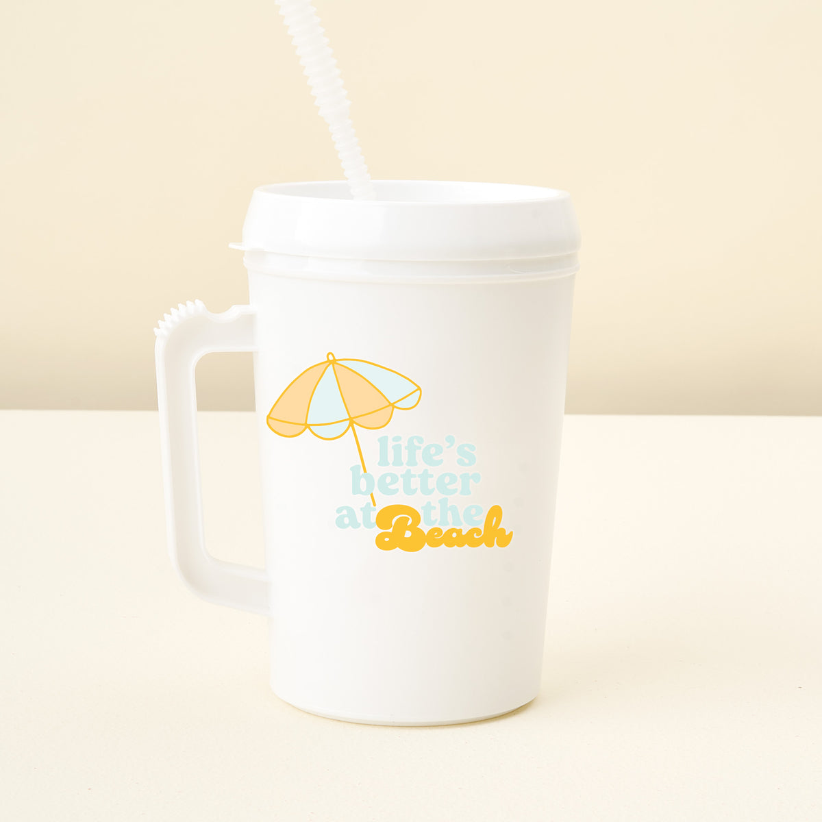 The Darling Effect Life’s Better At The Beach 34 oz Mega Mug is a white plastic mug with a handle and a straw lid, decorated with a yellow umbrella graphic and the phrase “Life’s better at the Beach” in blue and yellow lettering—a fun addition to any mega mug collection.