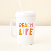 A white plastic Beach Life 34 oz Mega Mug by the-darling-effect, featuring a handle, straw, and vibrant “BEACH LIFE” lettering on the front, sits on a light surface against a beige background—ideal for keeping hydrated wherever you go.
