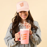 A woman in a denim jacket and pink hat holds the-darling-effect’s Mama Knows Best 34oz Mega Mug, sipping from a straw and showing off branded event drinkware against a beige background.