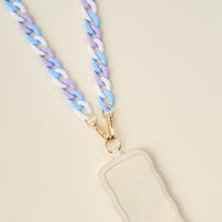 The Darling Effect's Tri Cool Hold the Phone Crossbody Chain features pastel blue, purple, and white links with gold clasps—an on-trend accessory for your cream, wavy-edged badge holder or phone, styled on a light beige surface.