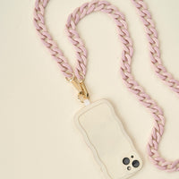 The Darling Effect's Duo Lilac Hold the Phone Crossbody Chain, featuring a pale pink chunky chain with gold clasps, is attached to a light phone case and displayed on a light beige surface.