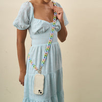 A woman in a light blue dress models The Darling Effect’s Tri Lively Hold the Phone Crossbody Chain, featuring a pastel multicolored strap and wavy-edged white holder, adding style against a beige backdrop.