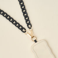 The Darling Effect's Midnight Black Hold the Phone Crossbody Chain features a wavy-edged, cream pendant with gold clasps on a black chain, shown against a light beige background.