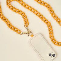 A smartphone with a beige wavy-edged case is attached to a thick, yellow crossbody chain strap with gold clasps, creating a fashionable accessory on a cream-colored surface.