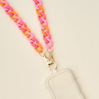 A close-up of The Darling Effect's Tri Sassy Hold the Phone Crossbody Chain, featuring chunky pink and orange links with gold clasps attached to a white item, set against a light beige background.