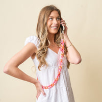 A smiling woman in a white dress holds a phone to her ear, showcasing The Darling Effect's Tri Sassy Hold the Phone Crossbody Chain—a bold pink and orange accessory—against a plain light background.