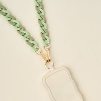 The Darling Effect’s Duo Sage Hold the Phone Crossbody Chain features a chunky light green and mint chain with gold clasps on a wavy cream card holder, creating a chic accessory set against a light beige background.