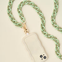 The Darling Effect's Duo Sage Hold the Phone Crossbody Chain, featuring chunky mint green and olive links with a gold clasp, is attached to a beige wavy-edged phone case resting on a light cream surface.