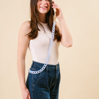 A smiling woman in a cream tank top and blue jeans holds her phone styled with the-darling-effect’s Marble Sky Blue Hold the Phone Crossbody Chain, featuring chic gold hardware.