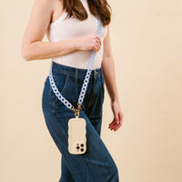 A person in a white sleeveless top and blue jeans holds a phone with a cream case, attached to the-darling-effect’s Marble Sky Blue Hold the Phone Crossbody Chain, worn crossbody style against a beige background.