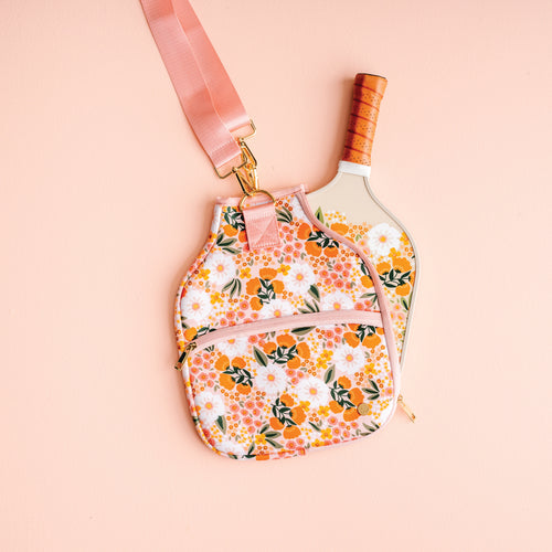The Darling Effect’s Sweet Meadow Pickleball Case and matching strap, shown on a light pink background with a white paddle, offer an elegant way to protect and carry your pickleball paddles.