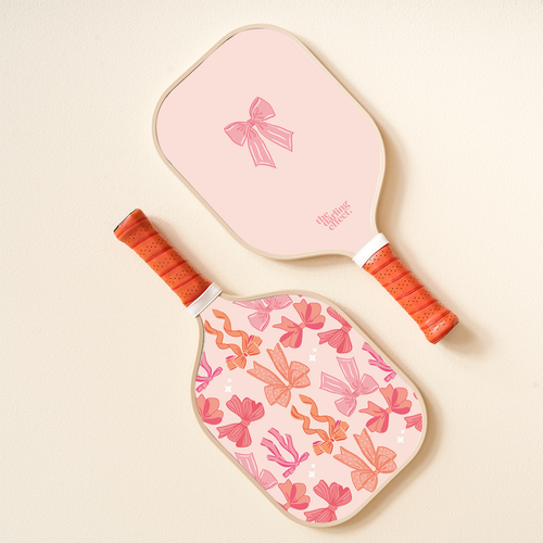 Elevate your pickleball game with the-darling-effect's Blushing Bows Pickleball Paddle, featuring orange grip handles and chic pink bow designs on a blush background.