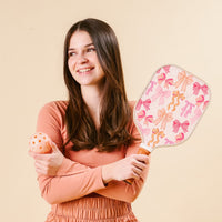 A young woman with long brown hair, wearing a peach top, smiles while holding the-darling-effect’s Blushing Bows Pickleball Paddle and an orange pickleball, ready for her next game against a beige background.