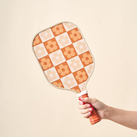 A hand with pink nails holds The Darling Effect's Flower Check Brown Pickleball Paddle, featuring an orange, white, and beige checkered floral design. Set against a light background, it's the perfect stylish choice for your next pickleball game.