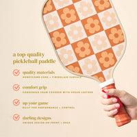 A hand holds The Darling Effect's Flower Check Brown Pickleball Paddle, with an orange and beige floral design. Quality materials, a comfort grip, and performance features offer a stylish edge for your pickleball game.