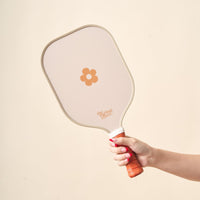 A hand with pink-painted nails holds The Darling Effect's Flower Check Brown Pickleball Paddle, featuring a brown flower design, against a plain beige background.
