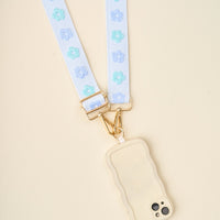 The Darling Effect's Blue Hold the Phone Crossbody Strap features a cream phone in a wavy-edged case, adorned with blue and light blue flowers and gold hardware. This stylish, hands-free accessory is shown resting on a light beige surface.