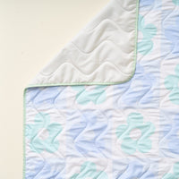 A close-up of The Darling Effect's Flower Check Blue On-the-Go Quilted Blanket, extra large with a pastel blue floral pattern, wavy stitching, light gray underside, and light green border—ideal for outdoor activities.