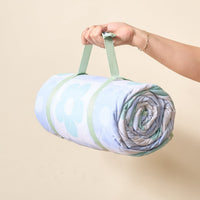 A hand holds The Darling Effect’s Flower Check Blue On-the-Go Quilted Blanket, featuring a green handle and a light blue quilt with large, light green and white flower patterns—ideal for outdoor activities.