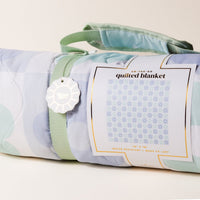 The Darling Effect’s Flower Check Blue On-the-Go Quilted Blanket features a light blue and pastel green geometric design, a matching strap and handle, plus “quilted blanket” tag—ideal for outdoor activities.