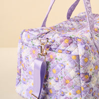 The Bouquet Beauty Purple On-the-Go Quilted Weekender Bag by the-darling-effect features a pastel floral design with daisies, gold hardware, and an adjustable strap—perfect as a charming travel bag.