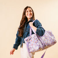 A woman with long brown hair in a denim jacket and white pants smiles while carrying the-darling-effect’s Bouquet Beauty Purple On-the-Go Quilted Weekender Bag with adjustable shoulder strap against a beige background.