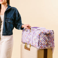 A woman in a denim jacket and white pants stands by a beige suitcase, with the-darling-effect's Bouquet Beauty Purple On-the-Go Quilted Weekender Bag featuring an adjustable strap placed on top, all set against a light background.