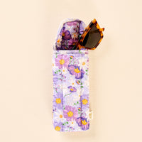A pair of tortoiseshell sunglasses is partially tucked into the-darling-effect’s Bouquet Beauty Purple Sunglasses Case—a soft, quilted case adorned with a floral pattern of purple and yellow flowers and featuring an easy-to-use magnetic flap closure—set against a light beige background.
