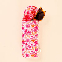 A pair of tortoiseshell sunglasses is partially tucked into the-darling-effect’s Paradise Palms Sunglasses Case, featuring a pink, orange, and red tropical palm tree print, set against a light beige background.