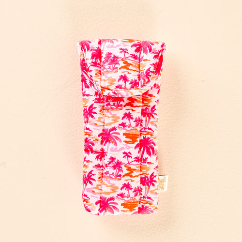 The Paradise Palms Sunglasses Case by the-darling-effect is a soft, quilted case featuring a pink, orange, and red palm tree pattern on a light background, shown against a pale peach surface.
