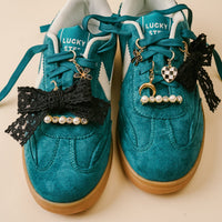 A pair of teal suede sneakers with Lucky Step insoles, styled with black lace bows, pearl beads, and the-darling-effect’s Midnight Moment Dazzle Shoe Charm Pack—featuring gold and silver moons, dice, and a flower—on the laces.