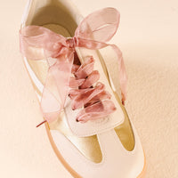 The Darling Effect's Sheer Mauve Step It Up Shoelaces—wide sheer pink ribbons tied in a large bow—adorn a white sneaker with gold accents, displayed on a light-colored textured surface.
