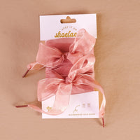 Sheer Mauve Step It Up Shoelaces by The Darling Effect, tied in bows on white "shoelaces" packaging labeled "Accessorize Your Shoes," rest on a light brown surface—ideal for adding a stylish finishing touch to your footwear.