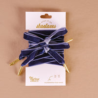 A pair of wide, stylish velvet blue shoelaces with gold tips are tied in bows and displayed on a branded card reading Velvet Blue Step It Up Shoelaces by The Darling Effect against a beige background.