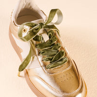 A metallic gold sneaker with cream accents and The Darling Effect Velvet Green Step It Up Shoelaces tied in a bow.