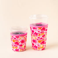 Two clear plastic cups, one small and one large, each fitted with a the-darling-effect Paradise Palms Sip Savvy Drink Sleeve featuring a vibrant pink, orange, and yellow palm tree pattern, are set against a plain light background.