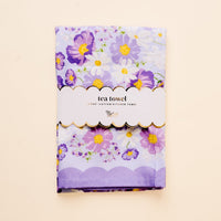 The-darling-effect Bouquet Beauty Purple Tea Towel is a 100% cotton kitchen towel with a floral pattern of purple, white, and yellow flowers on light lavender. It comes folded with a scalloped label reading "tea towel" and "100% cotton kitchen towel.