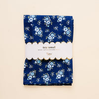The Endless Daydream Navy Tea Towel by the-darling-effect features small blue and white flowers, is folded with a scalloped “tea towel, 18x28” cotton kitchen towel label, and set against a light beige background.