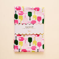 A folded cotton Pickleball Party Tea Towel by the-darling-effect features pink, yellow, and green checkered and striped bottle shapes, wrapped in a scalloped-edge white paper band labeled “tea towel.”.