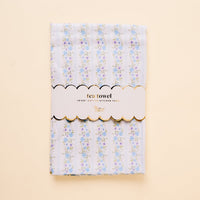 The-darling-effect’s Petal Parade Blue Tea Towel is a cotton kitchen towel (18x25) with light blue and white stripes, blue and pink floral accents, and a white label trimmed in gold that reads "tea towel" and "18x25 COTTON KITCHEN TOWEL.