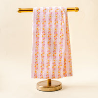 The Petal Parade Pink Tea Towel by the-darling-effect, featuring pink and orange florals, is draped over a gold stand against a light beige background.