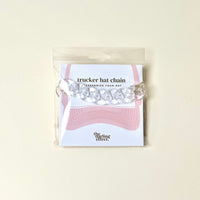 The Acrylic Tortoise Trucker Hat Chain by The Darling Effect is packaged on a card with a pink hat illustration and brand text, then sealed in a clear plastic bag against a plain background.