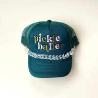 A dark green mesh trucker hat with "pickle baller" in colorful embroidery, featuring The Darling Effect's Acrylic Tortoise Trucker Hat Chain attached across the brim for a bold accessory.