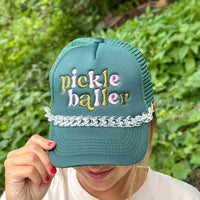 A person holds the brim of a green mesh trucker hat featuring colorful Pickle Baller embroidery and an Acrylic Tortoise Trucker Hat Chain by The Darling Effect. Green foliage is blurred in the background.