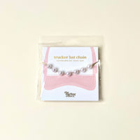 A Dainty Pearl-Gold Trucker Hat Chain by The Darling Effect is featured on a white background, accompanied by a pink hat illustration and the text “trucker hat chain” and “The Darling Effect” on the packaging card. This chic accessory pairs delicate pearls with subtle metallic gold chains, adding an elegant touch to your favorite trucker hat.