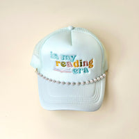 A light blue trucker hat with “in my reading era” embroidered in colorful letters and an open book design, finished with The Darling Effect’s Dainty Pearl-Gold Trucker Hat Chain—a chic metallic accessory draped across the front for added style.